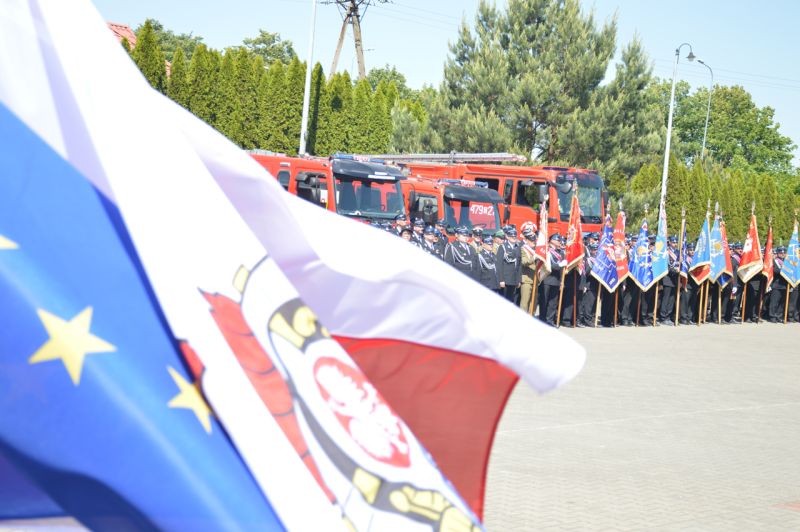 Zdjęcie przestawia flagi Polski, Unii Europejskiej oraz godła PSP w tle widać stojących strażaków